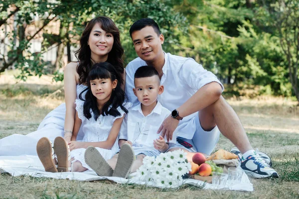 Portrait of an asian family in white clothes having picnic in a park, enjoying last warm days of the early fall. Parents and their children in a tree shadow.