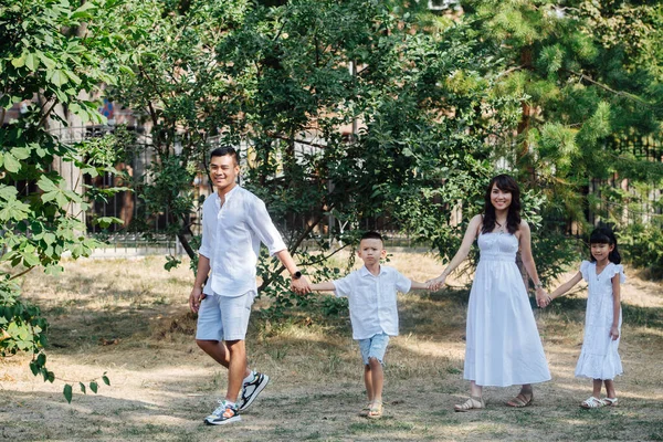 Joyful asian family in white clothes having a walk on a grass in a park, enjoying last warm days of the early fall. Slowly strolling between trees, holding hands.