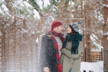 Parkta karlı kozalaklı ağaçların arasında birbirine yakın yürüyen mutlu çift. İkisi de şapka, ceket ve fular takıyor. Dallardan düşen kar taneciklerini fırlattı.