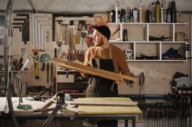 Büyük bir atölyede saat şapkalı bir marangoz elinde odun parçası tutuyor. Duvarda asılı araç sayısı.