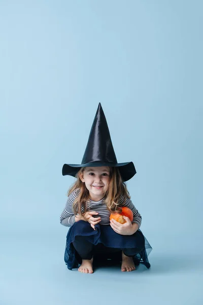 Ragazzina accovacciata in cappello da strega con piccola zucca, vestita per Halloween — Foto Stock