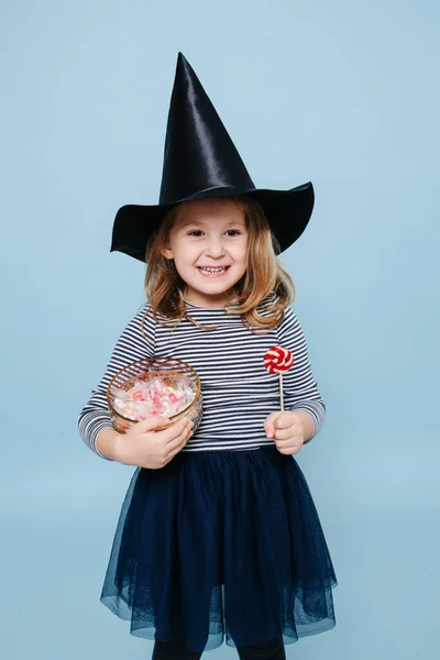 Adorabile ragazzina col cappello da strega che tiene caramelle e un lecca-lecca, orgogliosa di sé — Foto Stock