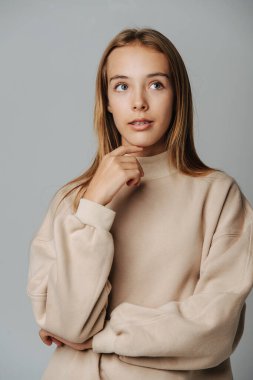 Hayalperest genç kız parantezlerle yukarı bakıyor, çenesine dokunuyor. Gri arka plana karşı. Sıcak bej bir uzun kolla.