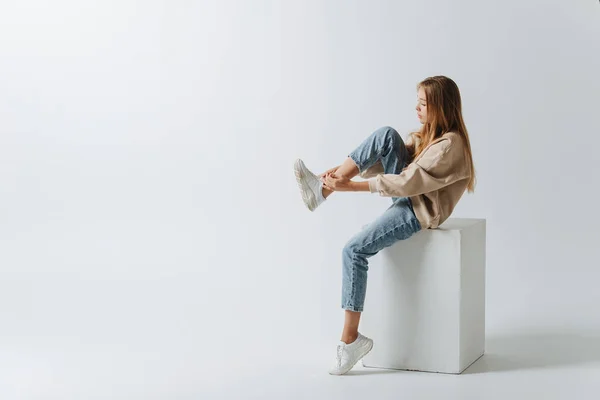 スニーカーを履いた10代の少女のロングショット 白い立方体に座っている 白い背景のスタジオで — ストック写真