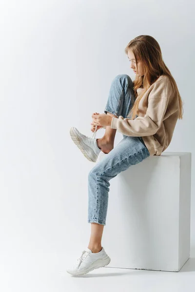 スニーカーを履いた10代の少女の横顔 白い立方体に座っている 白い背景のスタジオで — ストック写真