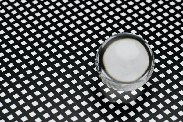 Clear glass toy sphere on a square holed black lattice. It distorts image like a lens. It makes square into a circle.