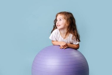 Yanaklarında gamzeleri olan mavi gözlü kıvırcık bebek leylak fitness topuna gülüyor.