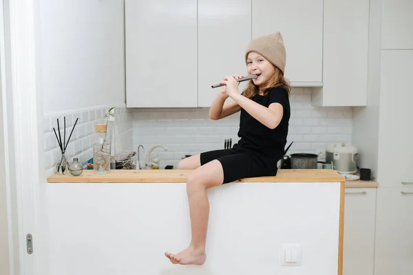 一个戴着米色帽子 拿着长笛玩耍的小女孩 坐在厨房的一张高高的桌子上 — 图库照片