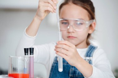 Bilim projesine odaklanmış bir kız, pipetin altına beyaz tuz tozu ile sıvı ekliyor. Yavaş ve kesin hareket etmeye çalışıyorum. Masanın arkasında. Ellerine odaklan..