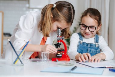 At kuyruklu kız mikroskoptan bakıyor, yanındaki notları alıyor. Kitaplar ve renkli sıvılarla dolu bir masanın arkasında..