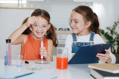 Hevesli küçük kızlar bilim projesi yapıyor, biri pipetten bir şişeye sıvı ekliyor, kimyasal bir reaksiyona neden oluyor, bol köpüklü bir gaz salıyor. İkisi de koruyucu gözlük takıyor..