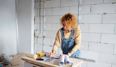Female construction engineer working on a blueprint on an improvised table, consulting with a phone app. Over a rough grey brick wall.