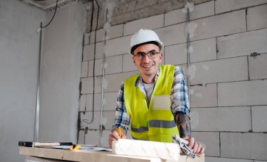 Cheerful construction engineer in white helmet drawing on a blueprint on an improvised board table. Over a rough grey brick wall.