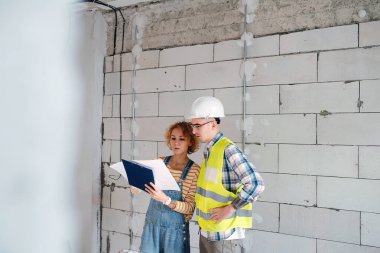 Serious looking engineering team going over a blueprint, discussing it. Next to a brick wall inside a building under construction.