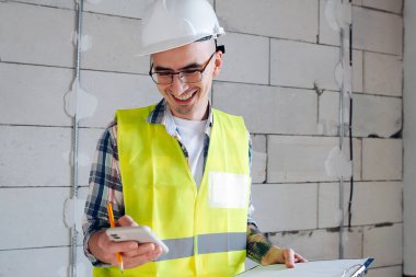 Happy construction engineer in white helmet looking at his phone, scrolling. Smiling widely. Inside an unfinished building