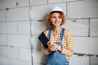 Happy female construction engineer in a white helmet holding a clipboard. Over a rough grey brick wall.