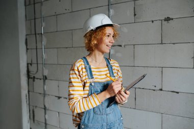 Social female construction engineer in a white helmet holding a clipboard, looking at someone. Over a rough grey brick wall.