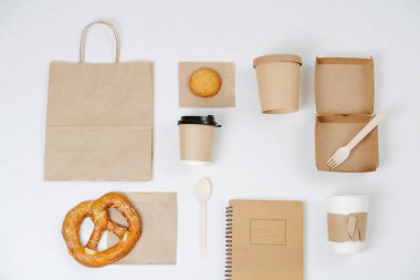 Paper eco friendly items arranged in composition. Carton compostable products over white. Cups, box, bag, cutlery, notepad completed with some pastry.
