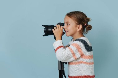 Çizgili süveter giyen küçük bir kız mavi arka planda tripoda monte edilmiş dijital bir kamerayla bakıyor..