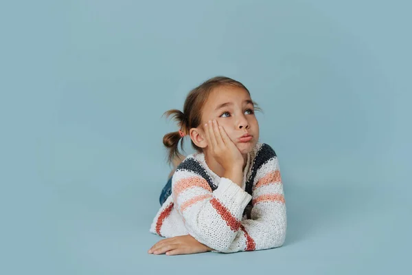 穿着条纹毛衣的小女孩躺在地板上的蓝色背景 靠着她的手 抬起头来 脸上露出忧郁的表情 — 图库照片
