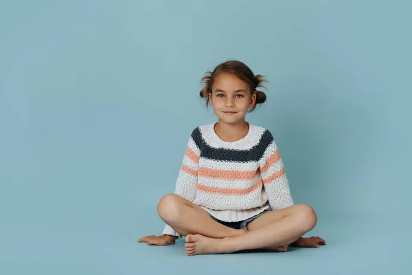 穿着条纹毛衣的漂亮小女孩双腿交叉坐在地板上蓝色背景 看着镜头 — 图库照片