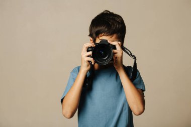 Hintli çocuk bej arka planda dijital kamerayla fotoğraf çekiyor. Yüzüne cihaz dayamışlar..