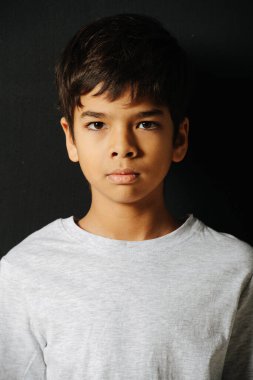 Siyah arka planda beyaz tişörtlü ciddi görünümlü Hintli çocuk. Dik dur, kameraya bak..