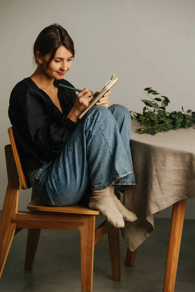 Happy woman sitting with legs up on a chair next to a round table, writing in a notepad. She\'s wearing mom jeans over black dress. Inside a room