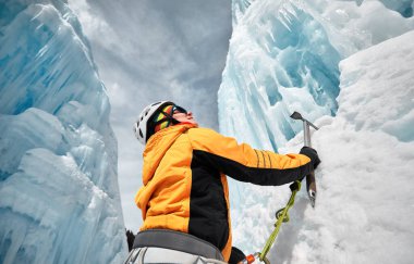 Kadın buz şelalesine tırmanıyor. Turuncu ceketli buz baltasıyla dağlarda.