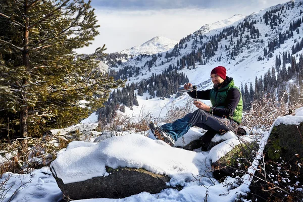 冬の山を背景に トウヒの森の魔法瓶から熱いお茶を注ぐ赤い帽子の髭を持つ男 — ストック写真