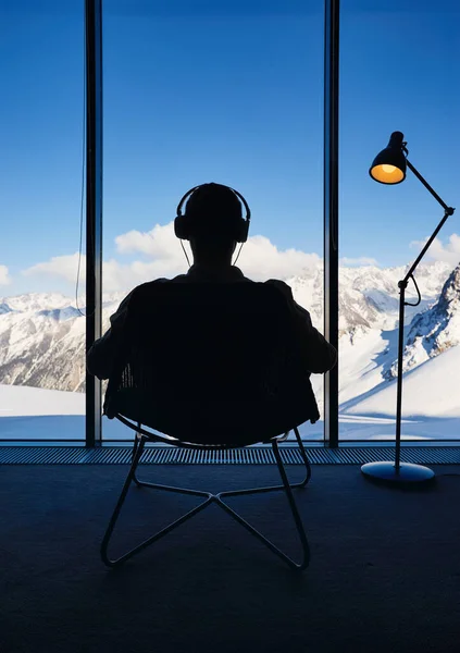 身披轮廓的男人坐在椅子上 用耳机在全景窗前听着音乐 平静地欣赏着美丽的雪山风景 酒店房间里的阁楼内行精明商人 — 图库照片