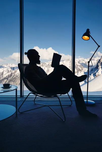 满头胡须的男子在椅子上的轮廓阅读着全景窗前的书 平静地观赏着美丽的雪山风景 酒店房间里的阁楼内行精明商人 — 图库照片