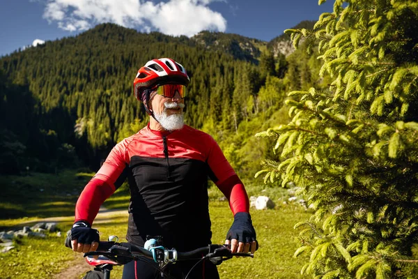 Portrait of Old cyclist with white beard holding handle bar of mountain bike in the green spruce forest in red costume in Almaty, Kazakhstan. Extreme Sport and outdoor recreation concept.