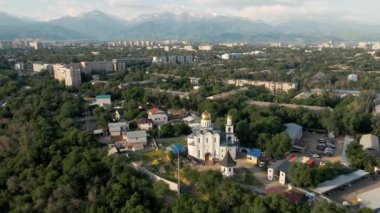 Aerial reveal shot Christian Church near the river with skyscrapers at Sairan lake with mountain view at sunrise in Almaty, Kazakhstan