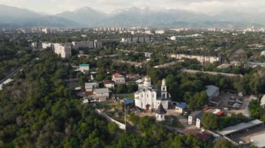 Aerial reveal shot Christian Church near the river with skyscrapers at Sairan lake with mountain view at sunrise in Almaty, Kazakhstan