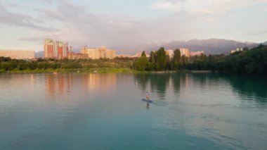 Man at stand up paddle boards SUP in the mountain lake Sairan in city Almaty in Kazakhstan. Drone shot aerial outdoor activity