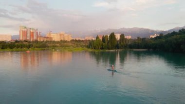 Man at stand up paddle boards SUP in the mountain lake Sairan in city Almaty in Kazakhstan. Drone shot aerial outdoor activity