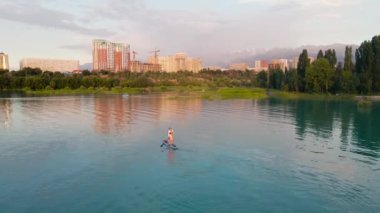 Man at stand up paddle boards SUP in the mountain lake Sairan in city Almaty in Kazakhstan. Drone shot aerial outdoor activity