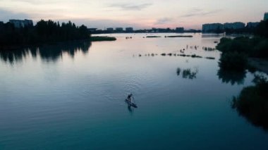 Man at stand up paddle boards SUP in the mountain lake Sairan at sunset in black silhouette in Almaty, Kazakhstan. Drone shot aerial outdoor activity