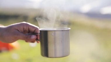 Iron Cup with hot beverage coffee with steam at slow motion at nature, brewing alternative coffee at camping. Making hot drink at picnic outdoors.
