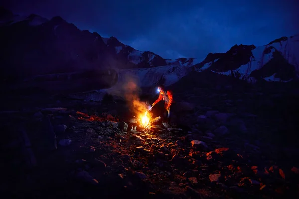 Tourist man in red jacket with headlamp near camp bonfire in the snow mountains under night sky with stars.