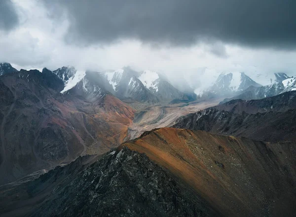 Aerial drone landscape shot of snow summit with ice glacier and and rocky wall at mountain valley with cloudy dark sky in Kazakhstan