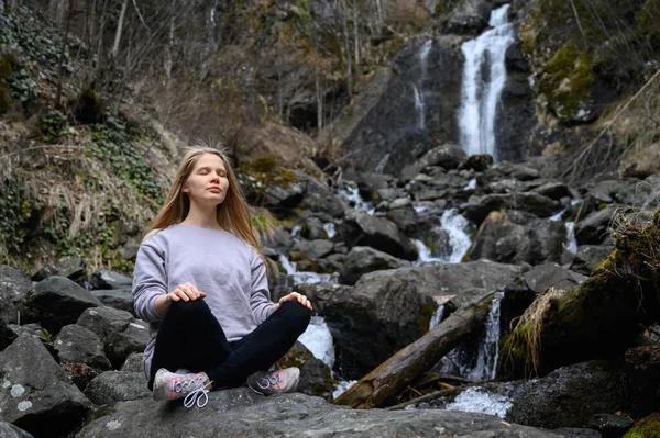 Menina senta-se em uma grande pedra e medita perto de uma cachoeira ao lado de um rio de montanha. Imagens De Bancos De Imagens Sem Royalties