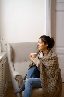 Zeki modaya uygun giyinmiş gülümseyen bayan elinde bir fincan çayla koltukta oturuyor. Gülümsemek, evde rahat bir atmosferde oturmak, güzel tasarım..