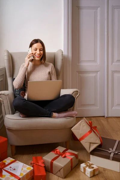 Mulher sentada em uma poltrona em casa, trabalhando no computador portátil, falando no celular — Fotografia de Stock