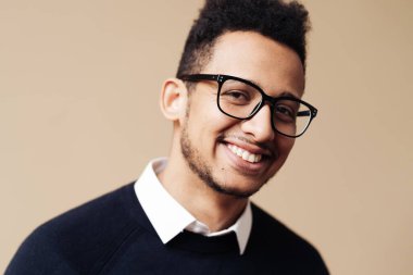 Siyah süveter ve gözlüklü, bej arka planda poz veren Afro yakışıklı bir adam.