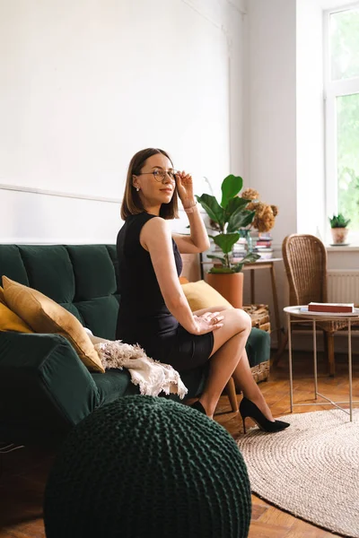 一位自信的女商人 身穿黑色经典礼服 戴着眼镜 坐在一个现代公寓的沙发上 有黄色枕头的绿色长沙发椅 — 图库照片