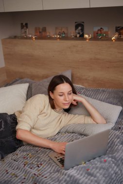 Gülümseyen genç bir kadın dizüstü bilgisayar kullanıyor, yatakta oturuyor.