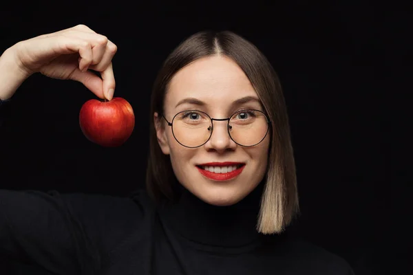 Χαμογελαστή γυναίκα που κοιτάζει την κάμερα κρατώντας ένα μήλο, φορώντας γυαλιά σε μαύρο φόντο — Φωτογραφία Αρχείου