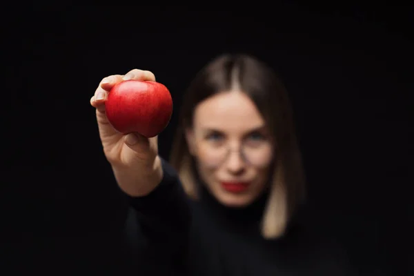 Χαμογελαστή γυναίκα δείχνει ένα μήλο στην κάμερα, φορώντας γυαλιά σε μαύρο φόντο — Φωτογραφία Αρχείου